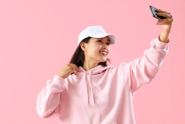 Beautiful young Asian woman in stylish hoodie taking selfie on pink background - Photo, Image