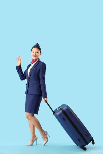 Όμορφη ασιατική αεροσυνοδός με βαλίτσα κουνώντας το χέρι σε μπλε φόντο - Φωτογραφία, εικόνα
