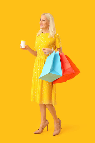 Mature woman with cup of coffee and shopping bags on yellow background - Photo, Image