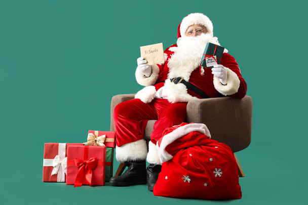 Santa Claus with letter, bag and Christmas gifts sitting on armchair against turquoise background - Photo, Image