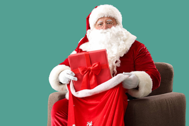 Santa Claus with bag and Christmas gift sitting on armchair against turquoise background - Photo, Image