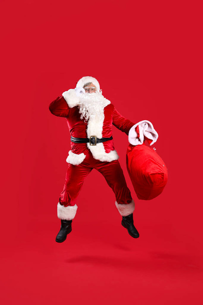Santa Claus con bolsa llena de regalos de Navidad saltando sobre fondo rojo - Foto, Imagen