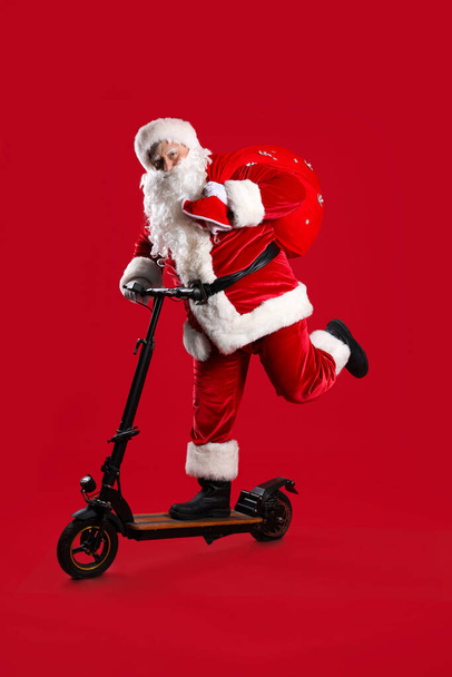 Santa Claus with bag full of Christmas gifts and kick scooter on red background - Photo, Image