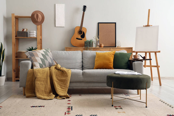 Interno del moderno soggiorno con divano grigio, pouf, cavalletto e chitarra acustica - Foto, immagini