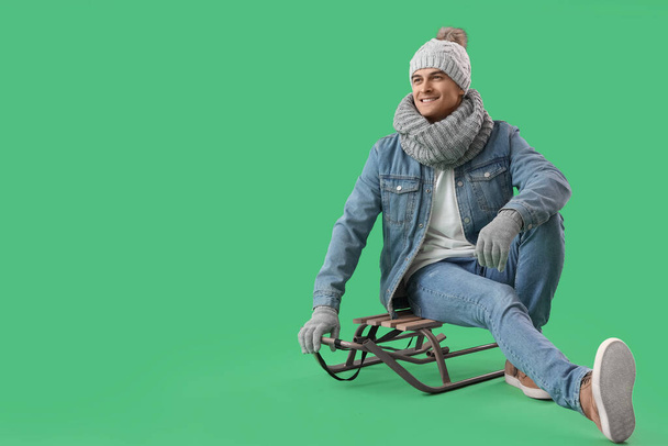 Schöner Mann in Winterkleidung sitzt auf Schlitten vor grünem Hintergrund - Foto, Bild