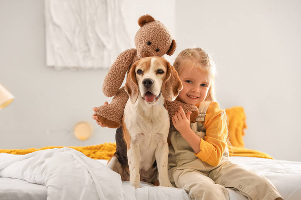 Nettes kleines Mädchen mit Spielzeugbär und Beagle-Hund im Schlafzimmer - Foto, Bild