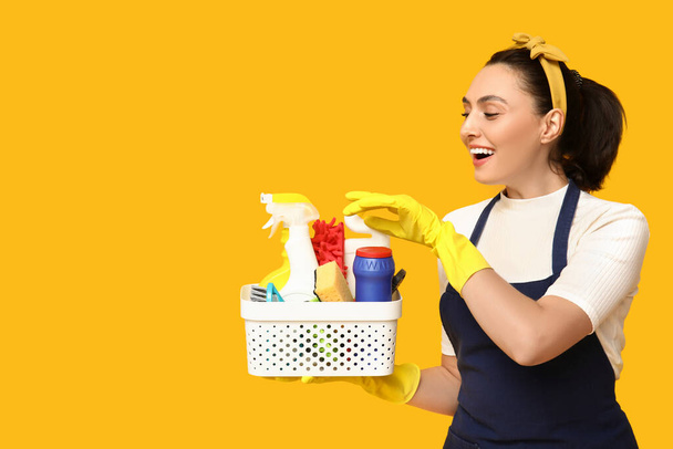 Portrait de jeune femme au foyer heureuse dans un tablier avec des fournitures de nettoyage sur fond jaune - Photo, image