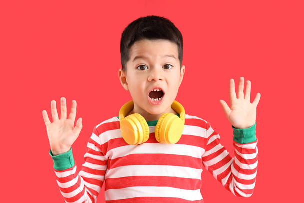 Schockierter kleiner asiatischer Junge mit Kopfhörern auf rotem Hintergrund - Foto, Bild