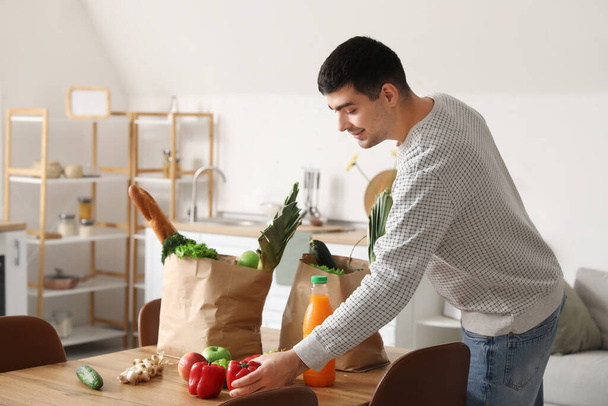 Junger Mann holt frisches Gemüse aus Einkaufstüten am Tisch in Küche - Foto, Bild