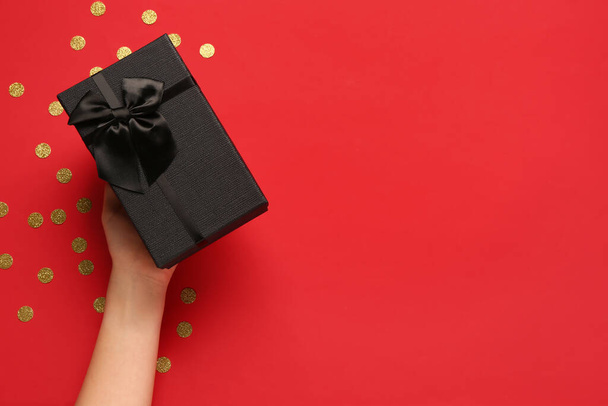 Mano femenina con caja regalo y confeti sobre fondo rojo. Viernes Negro - Foto, imagen