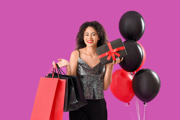 Schöne afroamerikanische Frau mit Einkaufstaschen, Geschenkbox und Luftballons auf lila Hintergrund. Black-Friday-Verkauf - Foto, Bild