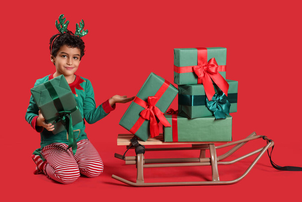 Cute little boy in elf costume pointing at gift boxes and sledge on red background - Photo, Image
