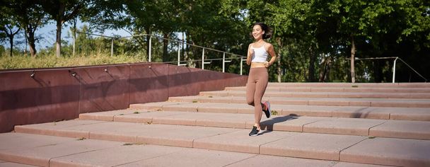 Chica de fitness saludable corriendo al aire libre en la calle, vistiendo uniforme, trotando en el aire libre y escuchando música en auriculares inalámbricos. - Foto, Imagen