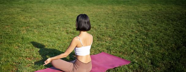Widok z tyłu sylwetki kobiety uprawiającej jogę, siedzącej na macie fitness i medytującej na zielonym trawniku. - Zdjęcie, obraz