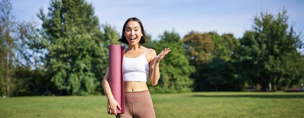 Aufgeregtes asiatisches Mädchen in Sportbekleidung, hält Gummimatte für Yoga, sieht überrascht und glücklich aus, steht im Park, trainiert an der frischen Luft. - Foto, Bild