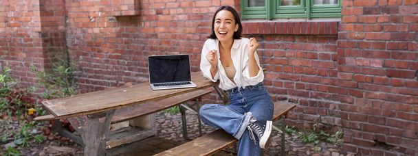 Disparo vertical de entusiasta chica asiática, se sienta con el ordenador portátil, la pantalla está vacía y en blanco, mujer riendo y celebrando, triunfando. - Foto, Imagen