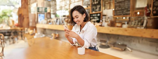Cafede oturan, kahve içen ve akıllı telefon kullanan Asyalı bir kadının dikey portresi. Yaşam tarzı ve insan kavramı - Fotoğraf, Görsel
