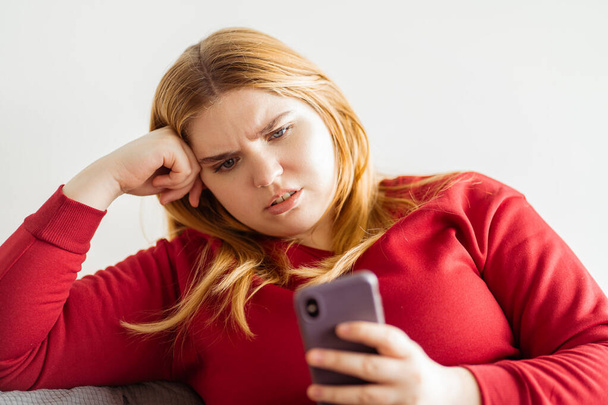 Κοντινό πλάνο πορτρέτο της γυναίκας συν μέγεθος κρατώντας μήνυμα κινητό τηλέφωνο συνομιλία, ενώ κάθεται στον καναπέ στο σαλόνι. Έννοια μέσων κοινωνικής δικτύωσης - Φωτογραφία, εικόνα