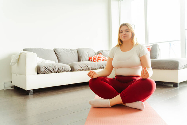Lächelnde schöne Frau mit geschlossenen Augen sitzt in Lotus-Pose auf einer Yogamatte zu Hause im Wohnzimmer. Konzept der Durchführung von Übungen, Training - Foto, Bild