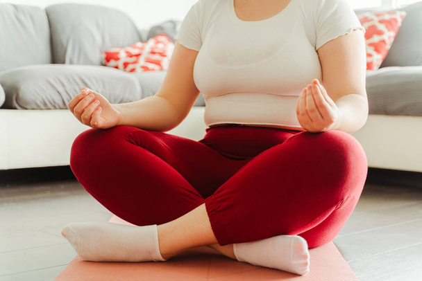 Closeup, selective focus, young woman sitting in lotus position on yoga mat, at home in living room. Concept of performing exercises, training - Photo, Image