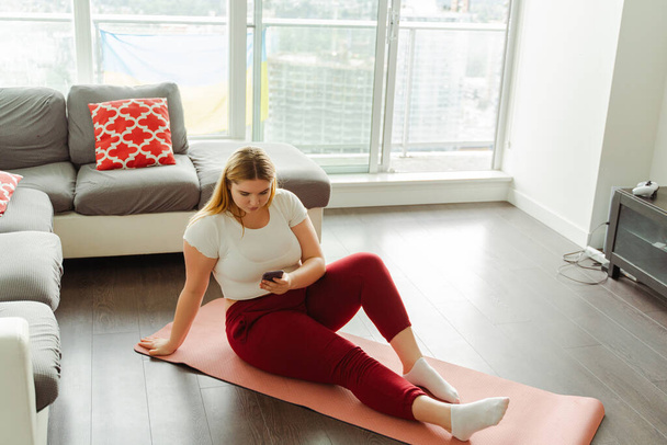 Linda joven mujer de tamaño grande sentada en una esterilla de yoga, sosteniendo el teléfono móvil, mensaje de texto, sentada en una esterilla de yoga, haciendo ejercicio en casa, relajándose. concepto de estilo de vida saludable - Foto, imagen