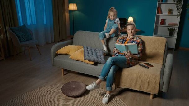 Little girl is bored near mom who is reading a book and not paying attention to her. Mom and daughter sitting on the couch in the living room - Фото, зображення