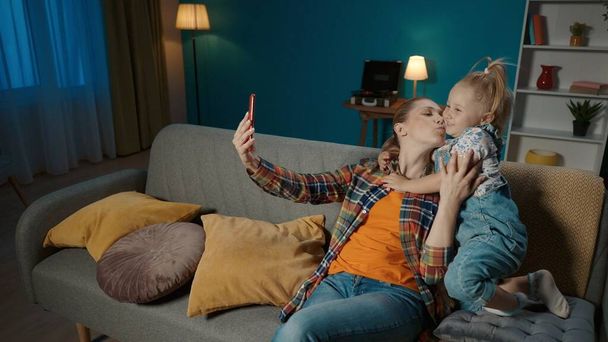 Happy mother and daughter taking selfies on smartphone while sitting on couch in living room at home. Cute little girl lovingly hugs her mom and mom kisses her daughter back - Fotografie, Obrázek