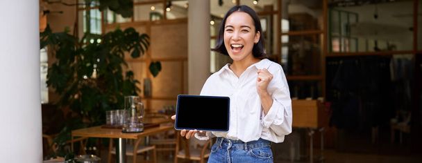 Smiling asian woman shows tablet screen and looks pleased, stands in cafe. - Photo, Image
