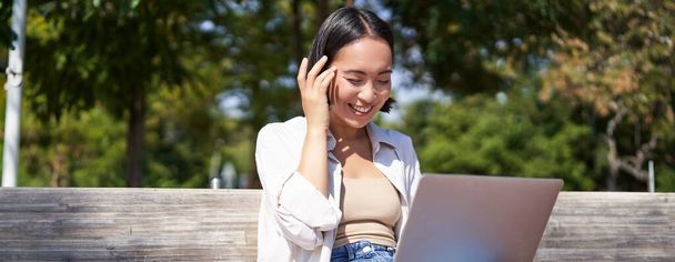 Όμορφο κορίτσι από την Ασία που γελάει, βλέποντας βίντεο στο laptop, ακούγοντας μουσική ή video chatting online ενώ κάθεται στο πάρκο στον πάγκο. - Φωτογραφία, εικόνα