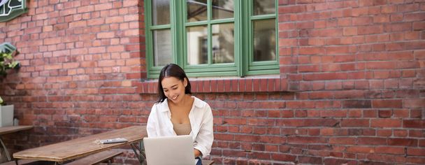 Vertical shot of asian woman working in outdoor cafe, sitting with laptop on bench and drinking coffee. Copy space - Photo, Image