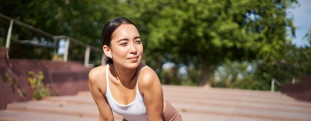Portrait de femme asiatique prenant une pause, respirant lourdement et haletant après avoir couru, jogger debout et essuyant la sueur sur le front, souriant heureux. - Photo, image