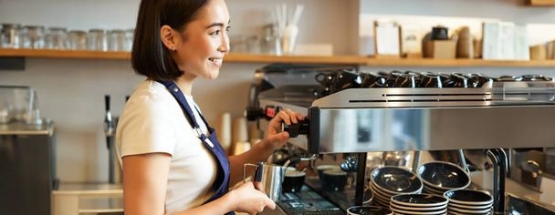 Souriant jeune fille asiatique barista, en utilisant la machine à café vapeur, lait fumant pour l'ordre cappuccino, debout dans le tablier, travaillant dans le café. - Photo, image