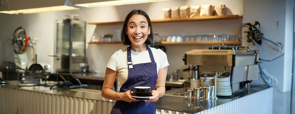 笑顔のアジアの女性バリスタの肖像、コーヒーを作る、お茶のカップを保持し、カフェクライアントにそれを取る、エプロンを身に着けて、カウンターの近くに立って. - 写真・画像
