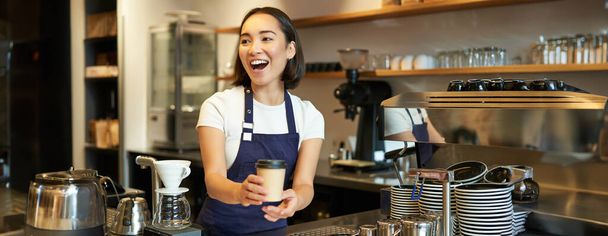 Portrait de fille asiatique souriante barista, donnant ordre dans le café, invitant l'invité à ramasser ordre à emporter près du comptoir, tenant à emporter tasse de café. - Photo, image