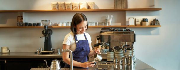 Happy lachende cafe eigenaar, meisje barista in schort, het maken van cappuccino, latte kunst met gestoomde melk, staan achter de toonbank. - Foto, afbeelding