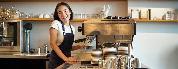 Chica barista sonriente feliz, camarero coreano en delantal, de pie cerca de la máquina de café, preparar el pedido para el cliente de la cafetería, mirando alegre a la cámara. - Foto, imagen