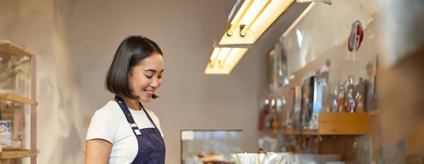 Pionowe ujęcie uśmiechniętej azjatki baristki, noszącej mundur, parzącej kawę, stojącej przy ladzie z filiżanką, pracującej w kawiarni. - Zdjęcie, obraz