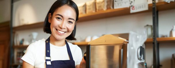 Вертикальний знімок бариста, азіатська дівчина, що готує молоко для капучино, готує лате для клієнта, одягнений у синій фартух, щасливо посміхається, працює в кафе
. - Фото, зображення