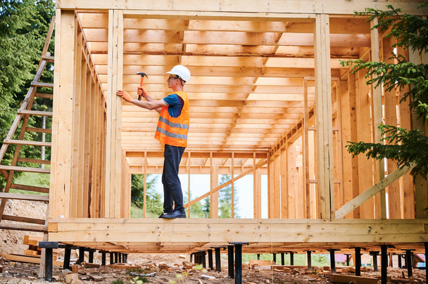 Wooden frame house being built by carpenter. Man wearing glasses hammering nails using hammer and is dressed in a protective helmet and a construction vest. Concept of modern ecological construction. - Foto, immagini