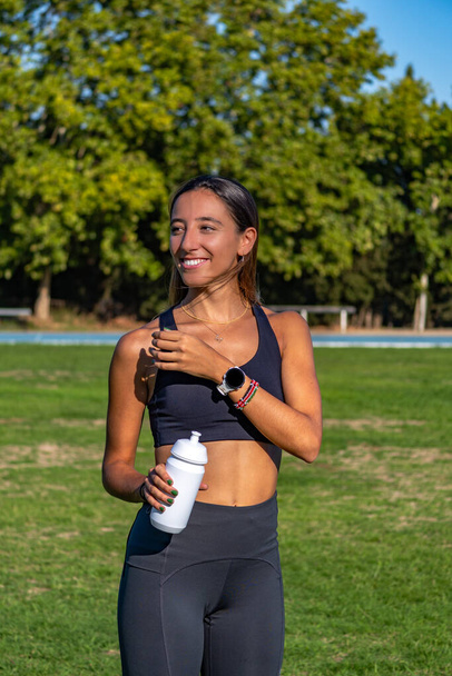 Beautiful slim young girl dressed in tight sportswear, holding with one hand an isotonic drink and smiling showing her white teeth looking at someone who makes her happy. - Photo, Image