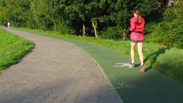 Parkta koşan sportif kadın sporu, spor rutini. Kırmızı elbiseli genç bir kız kolaylıkla koşar. Lastik kaplamalı yeşil koşu bandı. Güneşli sabah. - Video, Çekim