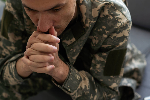 Vnitřní záběr nešťastného vojáka v maskáčové uniformě a s čepicí, jak drží ruku u obličeje, zatímco sedí na pohovce, přemýšlí a má zamyšlený, zamyšlený výraz ve tváři. Kvalitní fotografie - Fotografie, Obrázek