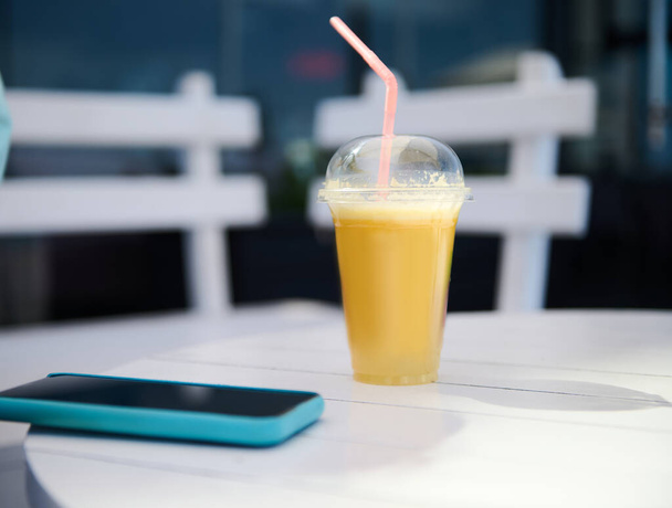 Um copo de suco de laranja saudável fresco e um telefone celular inteligente em uma mesa de madeira branca ao ar livre. Comida e bebida. Ainda vida - Foto, Imagem