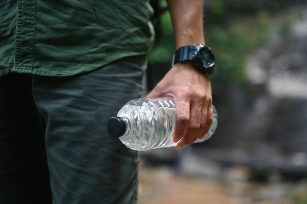 Colpo ritagliato di viaggiatore maschio in possesso di una bottiglia d'acqua con foresta offuscata su sfondo. - Foto, immagini