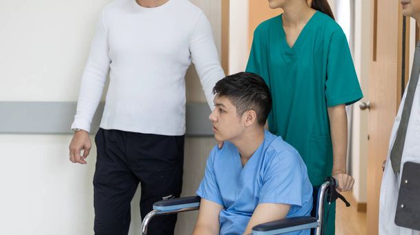 Jovem rapaz incapacitar paciente está sentado em uma cadeira de rodas sob o apoio de enfermeira e médico para se deslocar ao redor do corredor do hospital. - Foto, Imagem