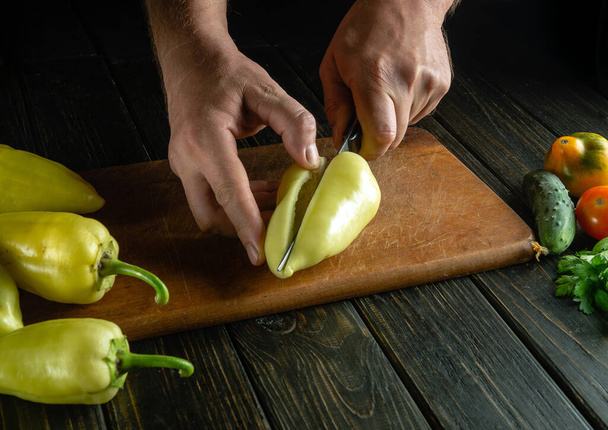 Cortar pimiento fresco amarillo con un cuchillo en una tabla de cortar para preparar un delicioso plato en casa. La idea de preparar una dieta vegetal en la mesa de la cocina con las manos de un chef. - Foto, Imagen
