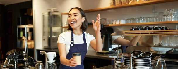 Портрет усміхненої азіатки-бариста, що видає замовлення в кафе, запрошуючи гостя забрати замовлення на винос біля лічильника, тримаючи чашку кави на винос
. - Фото, зображення