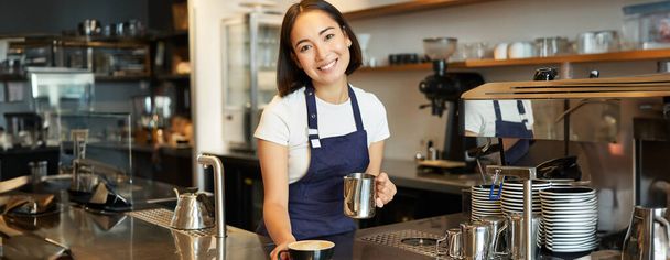 Hermosa joven barista femenina, haciendo capuchino, vertiendo leche al vapor para el arte del café con leche en la taza, dar su orden detrás del mostrador. - Foto, imagen