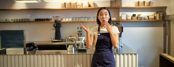 Kép izgatott ázsiai nő barista, néz meglepett, rámutatva smth kávézóban, álló mellett kávézó pult. - Fotó, kép