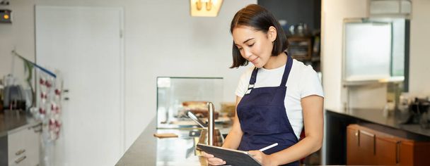 Pequenos cafés. Sorrindo asiático menina barista no avental, usando tablet como terminal de POS, processamento de ordem de café atrás do balcão. - Foto, Imagem